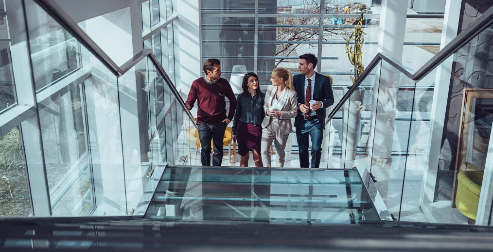 Kolleginnen und Kollegen im modernen Bürogebäude Vier Personen gehen gemeinsam eine gläserne Treppe hinunter und unterhalten sich in einem lichtdurchfluteten Bürogebäude.
