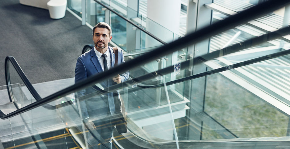 Geschäftsmann auf Rolltreppe in modernem Gebäude Ein Mann im Anzug fährt mit einer Rolltreppe in einem lichtdurchfluteten Bürogebäude nach oben und blickt konzentriert nach vorn.