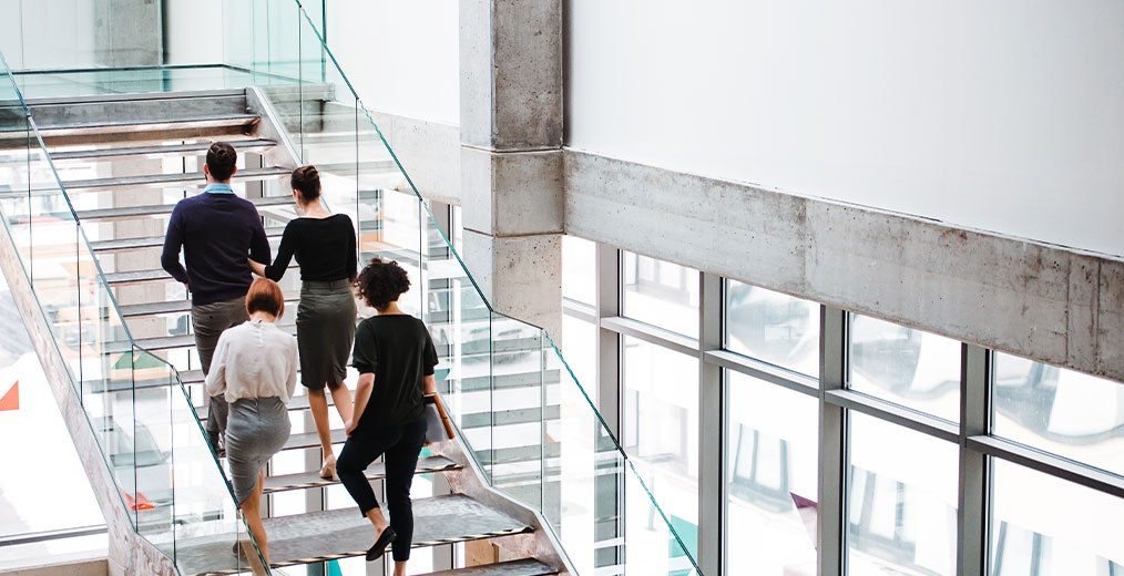 Vier Personen in Businesskleidung steigen eine gläserne Treppe in einem hellen, modernen Bürogebäude hinauf.
