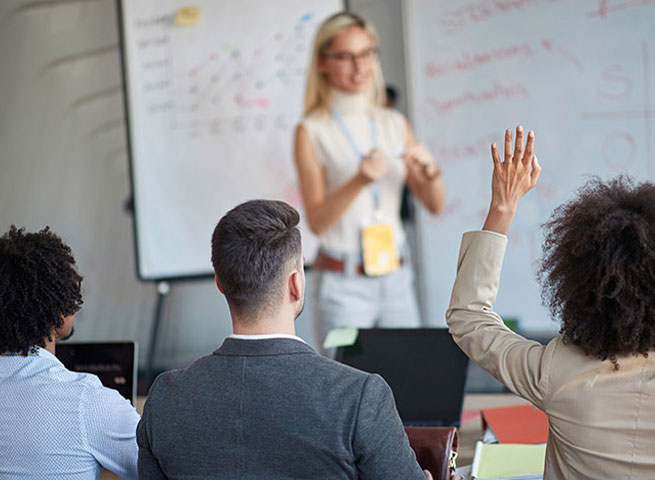 Eine Frau steht vor einem Whiteboard und erklärt etwas einer Gruppe von Teilnehmenden.