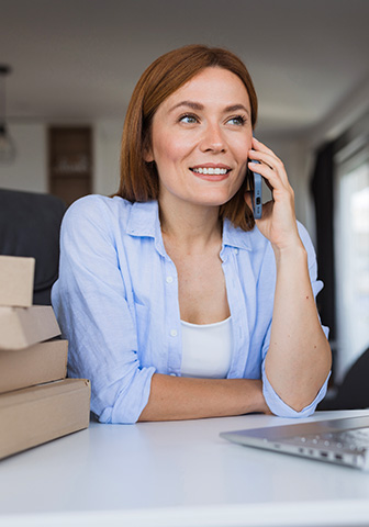 Frau telefoniert am Schreibtisch mit Laptop und Paketen Lächelnde Frau führt ein Telefonat am Laptop, neben ihr liegen mehrere Kartons.