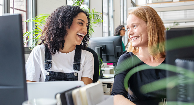 Zwei Kolleginnen lachen gemeinsam im Büro Zwei Frauen sitzen nebeneinander an Schreibtischen und unterhalten sich fröhlich.