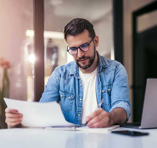 Mann arbeitet konzentriert an Dokumenten im Büro Nachdenklicher Mann mit Brille schreibt und prüft Unterlagen am Schreibtisch.
