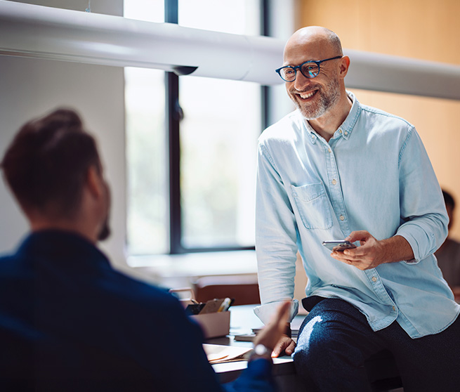 Lächelnder Mann sitzt auf Schreibtisch und spricht mit Kollegen Entspannter Mann mit Brille unterhält sich freundlich mit einem Kollegen im Büro.