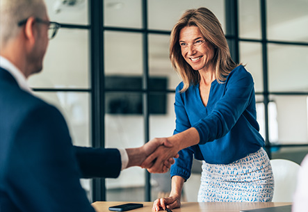 Frau begrüßt Geschäftspartner mit Handschlag im Büro Lächelnde Frau in blauer Bluse schüttelt einem Kollegen im Besprechungsraum die Hand.
