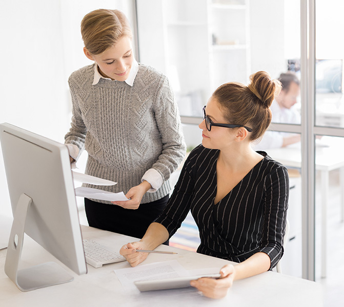 Zwei Mitarbeiterinnen im Büro arbeiten gemeinsam am Computer und besprechen Unterlagen.