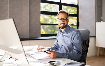 Buchhalter in Bielefeld eschäftsmann mit Brille arbeitet lächelnd am Schreibtisch mit Laptop und Unterlagen.