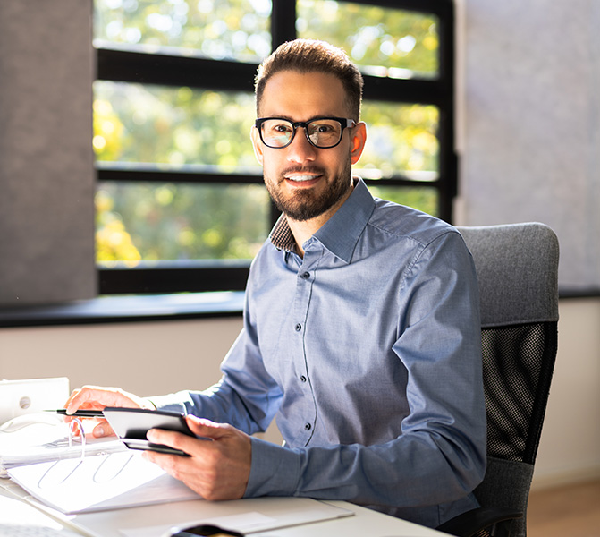 Buchhalter-Jobs in Bielefeld Geschäftsmann mit Brille sitzt am Schreibtisch und arbeitet mit Tablet und Unterlagen.