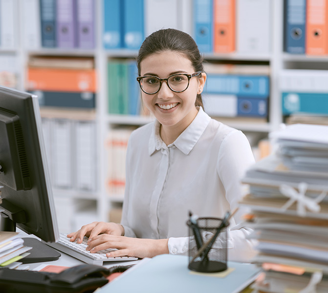 Sachbearbeiterin bei der Arbeit im Büro in Frankfurt Eine junge Frau mit Brille sitzt an einem Schreibtisch im Büro, tippt auf einer Tastatur und lächelt freundlich in die Kamera. Im Hintergrund sind bunte Aktenordner in Regalen zu sehen.