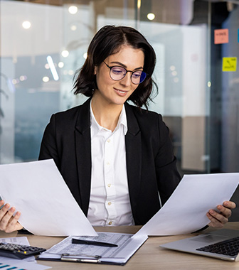 Frau im Büro mit Laptop und Papieren überprüft Unterlagen.