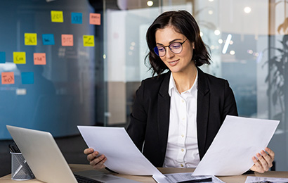 Steuerfachangestellter in Bielefeld Geschäftsfrau mit Brille prüft Unterlagen am Schreibtisch mit Laptop und Taschenrechner.