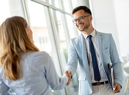 Geschäftliches Händeschütteln Zwei Personen begrüßen sich mit einem Handschlag in einem hellen Büro, im Hintergrund arbeitet eine weitere Person.