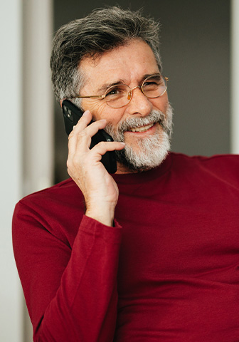 Terminvereinbarung in Essen Älterer Mann mit Brille und rotem Pullover telefoniert lächelnd im Büro, mit Notizen und Laptop auf dem Tisch.
