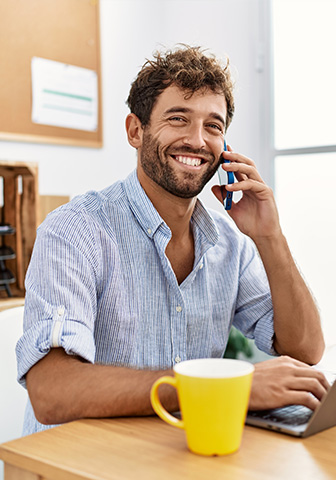 Mann telefoniert und lächelt am Schreibtisch mit Laptop. Ein junger Mann im gestreiften Hemd sitzt am Tisch, telefoniert und lächelt. Neben ihm liegt ein Laptop.