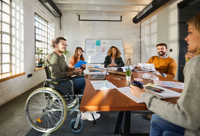 Eine Gruppe von fünf Personen, darunter eine Person im Rollstuhl, arbeitet gemeinsam an einem Tisch.