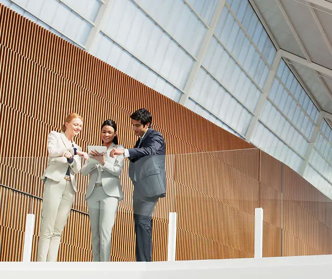 Drei Personen im Business-Outfit stehen in einem modernen Geb&auml;ude und sehen sich ein Tablet an.
