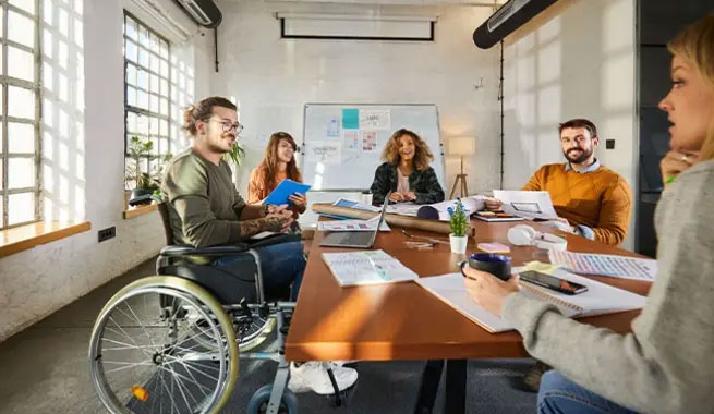 Eine Gruppe von f&uuml;nf Personen, darunter eine Person im Rollstuhl, arbeitet gemeinsam an einem Tisch.