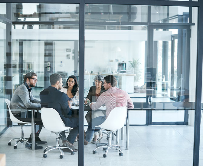 Fünf Personen sitzen an einem runden Tisch und führen eine Besprechung in einem verglasten Büro.