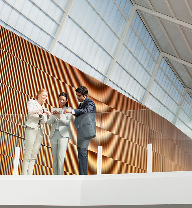 Drei Personen im Business-Outfit stehen in einem modernen Gebäude und sehen sich ein Tablet an.