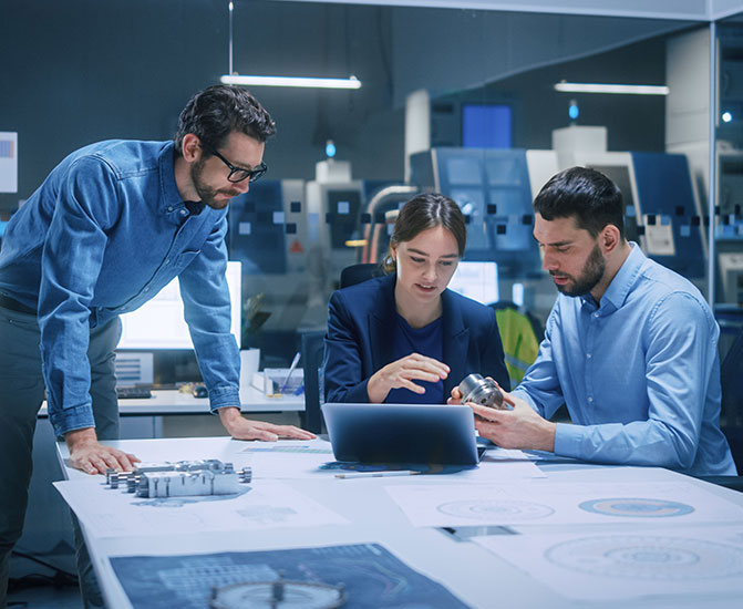 Drei Personen besprechen technische Zeichnungen und Bauteile an einem Tisch im Büro.