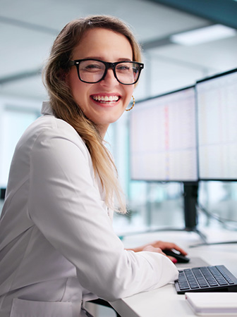 Fachwissen und Marktkenntnis Analystin lächelt am Arbeitsplatz vor mehreren Monitoren mit Daten- und Tabellenansichten.