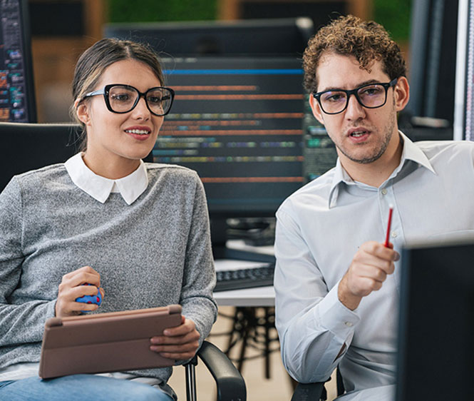 Mehrwert bei der Besetzung von IT-Positionen Zwei IT-Fachkräfte sitzen gemeinsam vor Monitoren mit Code und besprechen Programmierlösungen.