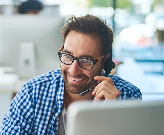 Ein Mann mit Brille und kariertem Hemd sitzt in einem modernen Büro, trägt ein Headset und schaut lächelnd auf den Bildschirm, während er offenbar ein Gespräch führt oder jemandem am Computer hilft.