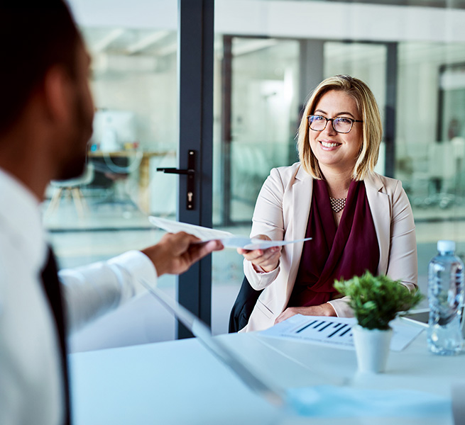 Lächelnde Frau reicht einem Kollegen ein Blatt Papier in einem modernen Besprechungsraum.