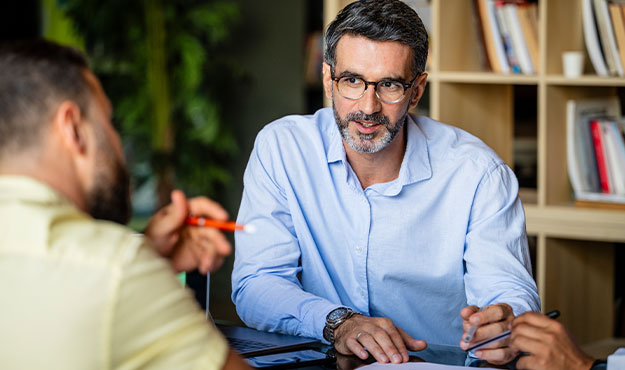 Zwei M&auml;nner im pers&ouml;nlichen Gespr&auml;ch am Tisch. Der Berater h&ouml;rt aufmerksam zu und bespricht gemeinsam mit seinem Gespr&auml;chspartner wichtige Themen.