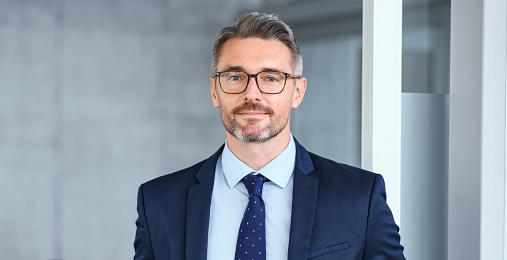 Geschäftsmann im Anzug steht im Büro und lächelt Selbstbewusster Mann in Anzug und Krawatte steht in modernem Büro und blickt in die Kamera.