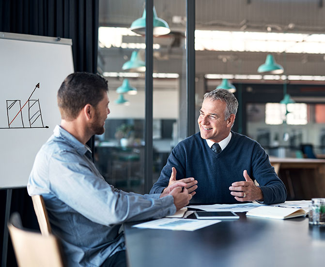 Geschäftsgespräch zwischen zwei Kollegen Zwei Männer sitzen an einem Konferenztisch und führen ein positives, fachliches Gespräch, während im Hintergrund eine Grafik mit wachsendem Diagramm zu sehen ist – Symbol für Unternehmenswachstum und Zusammenarbeit.